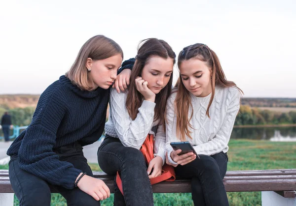 Three girls schoolgirls teenagers 13-15 years old, autumn day summer city park, holding smartphone, watching video app social networks Internet, casual sweater clothes relaxing after school vacation.