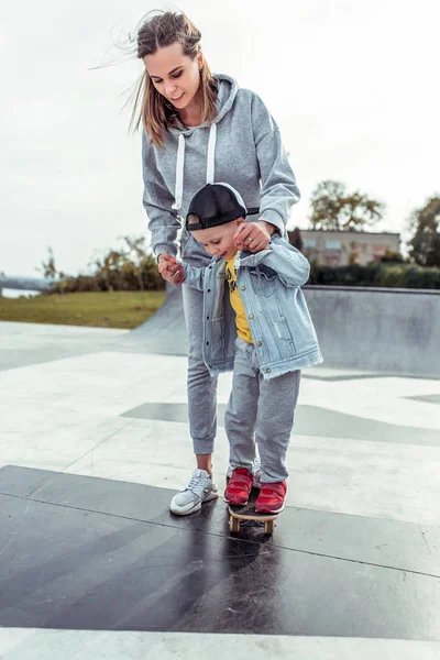 Frau Mutter lehrt Skateboarden, unterstützt Trainingspflege, kleiner Junge Sohn, trainiert Sommer Stadt lernt Skateboard, Herbst Sportplatz, glücklich lächelnd Freizeitkleidung. — Stockfoto