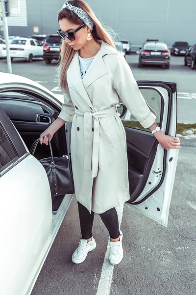 Menina bonita na capa de chuva, na cidade de outono de verão, sai e entra no carro interior segurando saco com compras da loja. Aluguel de carros, partilha de carros. Estacionamento no shopping . — Fotografia de Stock