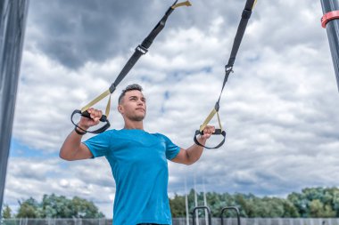 Erkek sporcu, kendini menteşelere çekiyor, elastik bantlarla şınav çekiyor, yazın şehirde. Gençlik yaşam tarzı spor motivasyonu. Metni kopyalamak için boş alan. Spor kıyafetleri tişörtü.