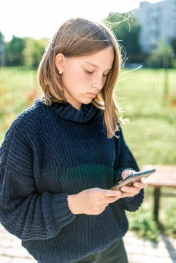 Genç kız 14-15 yaşlarında, sonbahar günü, sokak şehrinde duruyor, akıllı telefonunu tutuyor, internete bir başvuru formu okuyor ve yazıyor. Sosyal ağlar. Günlük kalın kazaklar..