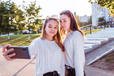 İki genç kız telefonda fotoğraf çekiyor, sonbahar günü şehirde, cep telefonlarına, rahat kıyafetlere, kazaklara, okuldan sonra yürümeye bakıyorlar. Akıllı telefondaki selfie..