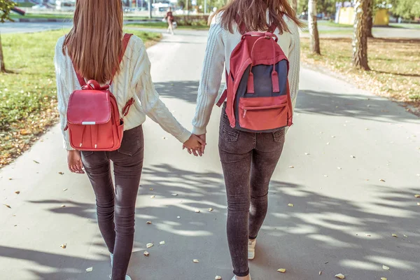 Две девочки-подростки ходят по городу осенним днем летом, держатся за руки, оглядываются назад, повседневная одежда, джинсы и свитер, гуляют после школы и колледжа. Рюкзаки с учебниками и тезисами . — стоковое фото