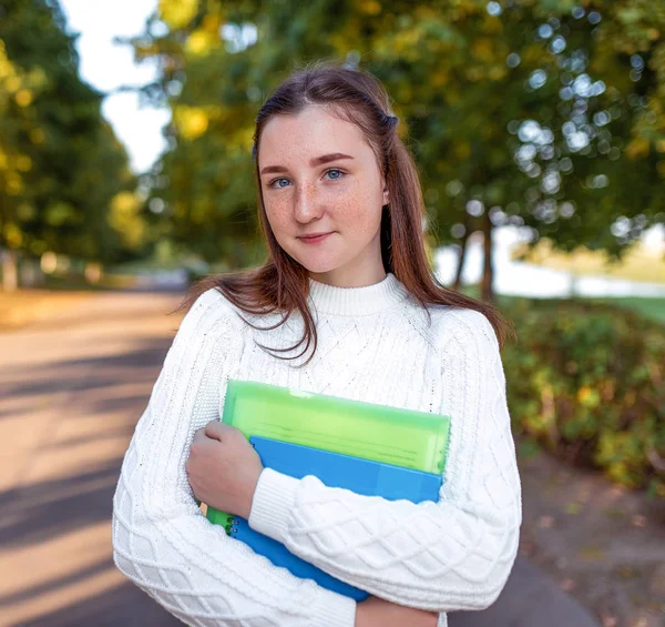 Счастливая девочка-подросток улыбается, стоя в летнем парке, на фоне осенних деревьев. В руках тетрадь учебники и заметки, подготовка к школе колледжа. Тёплый свитер . — стоковое фото