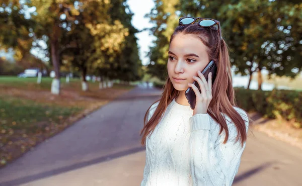 Gadis remaja membuat panggilan telepon, di musim panas di taman, latar belakang pohon musim gugur, mendengarkan pesan suara, online di aplikasi internet, ruang kosong untuk teks salinan. Baju hangat sweater putih . — Stok Foto