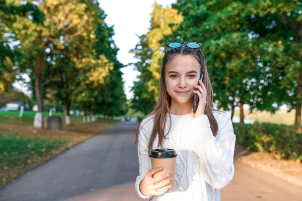 Teenager-Mädchen telefoniert, Sommerpark, Hintergrund Herbst Bäume, hört Sprachnachricht, Online-Internet-Anwendung, Freiraum Text kopieren. warme Kleidung weißer Pullover. hält Tasse Kaffee Tee Hand. — Stockfoto