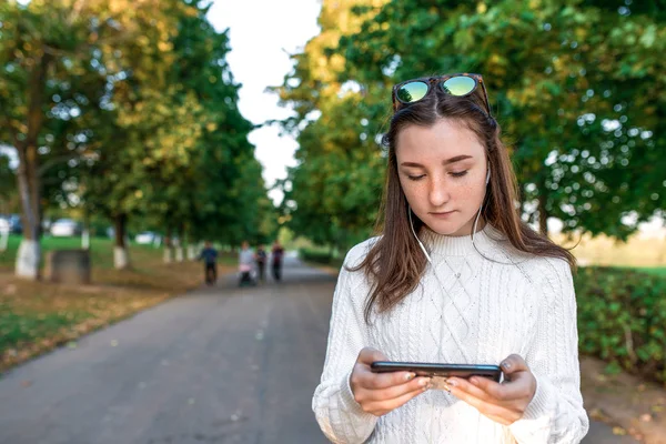 teenager girl watches video on phone, summer park, background autumn trees, listens to music with headphones, online in Internet application, free space for copy text. Warm clothes white sweater.