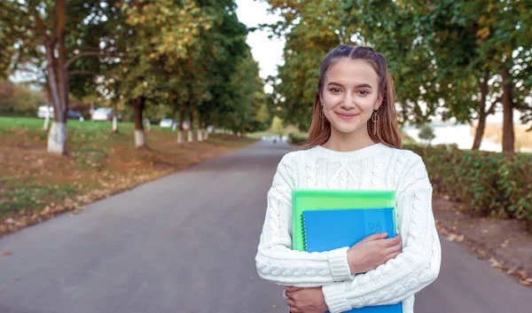 Heureuse adolescente souriante joyeuse joyeuse, se tient parc d'été arbres d'automne fond, espace libre pour copier le texte. Dans les mains notes de cahier de notes de manuels, école de préparation collège. Pull chaud pour vêtements . — Photo