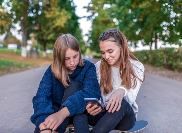 2人の女の子のガールフレンド、幸せな笑顔に座って、学校の後に10代、スケートボードをリラックスし、都市、喜びを楽しんでいます。手のスマートフォンでは、オンラインアプリのソーシャルネットワークでビデオを見て、カジュアルな服. — ストック写真
