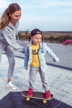 Genç kadın anne kaykay öğretmeni, küçük oğlan, kaykay. Hafta sonu parkta dinlenin. Her gün kalın giysiler, sonbahar günü. Mutlu gülüşler gülüşler ve neşe.