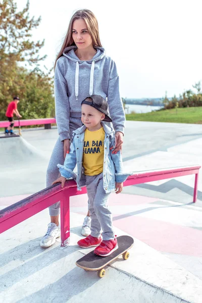 Mladá žena maminka učí skateboarding, malý syn chlapec, skateboard. Odpočiňte si o víkendu na podzim na ulici v parku. Každý den teplé oblečení, podzimní den. Podpora péče a výchovy. — Stock fotografie