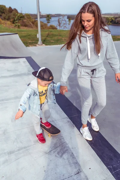 Genç kadın anne kaykay öğretmeni, küçük oğlan, kaykay. Hafta sonları spor sahasında dinlen. Her gün kalın giysiler, sonbahar günü. Destek ve yetiştirme. — Stok fotoğraf