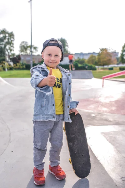 Liten pojke 3-5 år gammal, lära sig rida skateboard, höst dag, casual varma kläder. Denim med basebollkeps. Körlektioner, första erfarenhet, utomhussport. Glad leende, hand gest finger upp. — Stockfoto