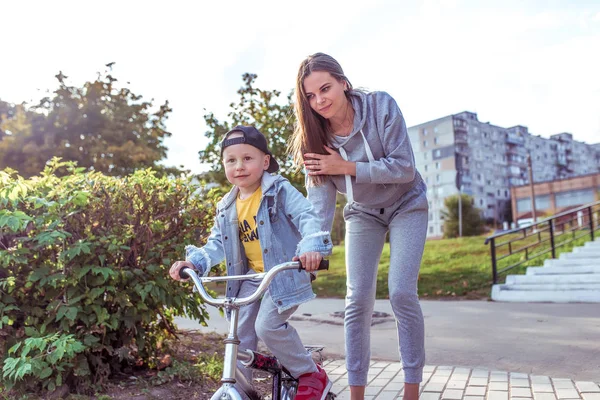 Genç kadın anne çocuğa bisiklet sürmeyi öğretiyor, küçük oğlan. Sonbaharda parkta dinlen. Her gün kalın giysiler, sonbahar günü. Ebeveyn bakımını ve yetiştirilmeyi destekle. — Stok fotoğraf