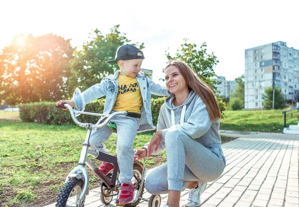 Молодая женщина мама учит детей ездить на велосипеде, маленький мальчик сын. Отдых в выходные осенью в парке. Каждый день теплая одежда, осенний день. Поддержка родительского ухода и воспитания. Счастливый смех, улыбка и радость . — стоковое фото