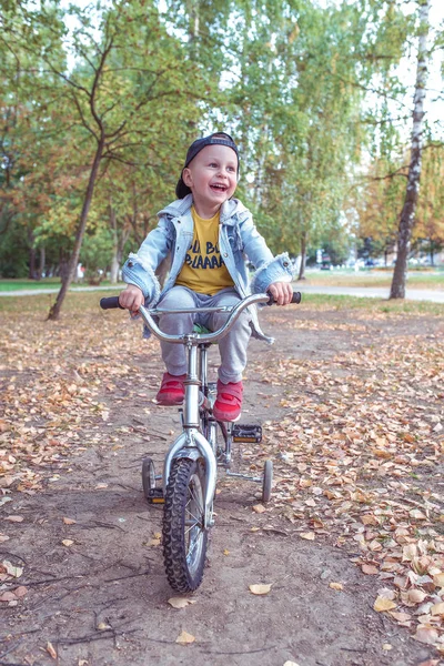 Anak laki-laki berusia 3-5 tahun, belajar mengendarai sepeda, hari musim gugur, pakaian hangat biasa. Denim dengan topi baseball. Belajar mengemudi, pengalaman pertama, olahraga di taman. Senyum bahagia tertawa dan bersukacita, bersenang-senang . — Stok Foto