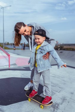 Genç anne oğluna ata binmeyi öğretir, destek sevgi ve şefkattir, 3-5 yaşındaki mutlu küçük çocuk, yaz sporları şehri, kaykay sürmeyi öğrenir, duygu, neşe, keyif, rahatlama keyfi.