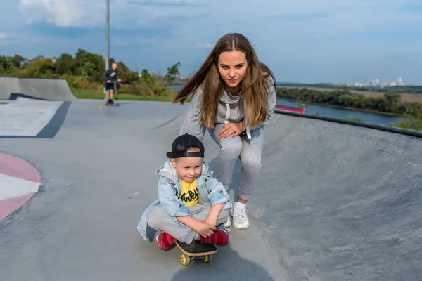 Perempuan ibu naik dan membantu anak-anak di skateboard, anak kecil 3-5 tahun, musim panas di lapangan olahraga di kota, musim gugur, belajar untuk naik skateboard, emosi cinta, perawatan, relaksasi dan menikmati . — Stok Foto