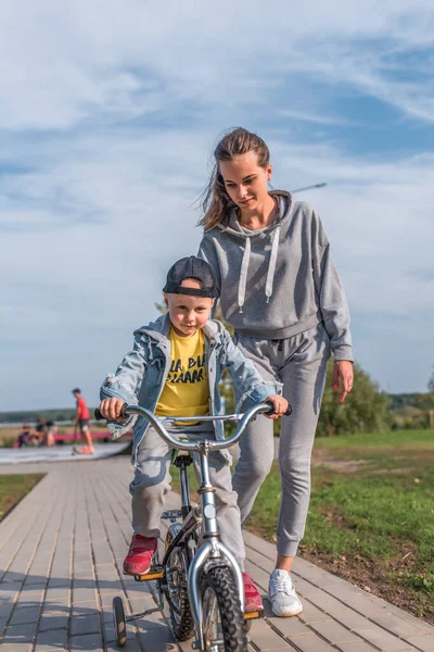 Kadın anne bisiklete binen çocuğa bakar ve yardım eder, 3-5 yaşındaki küçük çocuk, yaz kenti, sonbahar bisiklet sürmeyi öğrenir, sevgi, ilgi, rahatlama ve keyif. Günlük kıyafet, kot pantolon.. — Stok fotoğraf