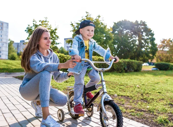 Kadın anne çocuk bisikletini destekler ve yardım eder, neşeli insanlar sevinir ve güler, 3-5 yaşındaki küçük çocuk, yaz kenti, sonbahar, bisiklet sürmeyi öğrenir, sevgi, ilgi, rahatlama ve keyif alma duyguları. — Stok fotoğraf
