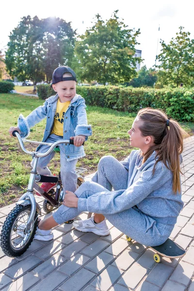 Ibu wanita membantu anak-anak bersepeda, bergembira gembira, anak kecil berusia 3-5 tahun, kota musim panas, musim gugur, belajar mengendarai sepeda, emosi cinta, kasih sayang, santai. Skateboard duduk . — Stok Foto