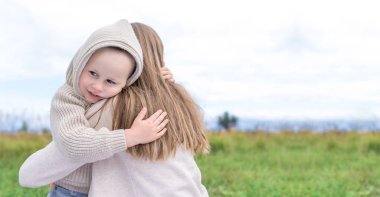 Küçük oğlan çocuğu annesine mutlu, şefkatli bir şekilde sarılıyor. Sonbahar günü çocuk bakımı, sıcak kıyafetler, yaz doğası. Arkadan bakıyorlar, rahatlıyorlar, duyguları sevindiriyorlar, şefkati seviyorlar. Metin için boş alan.