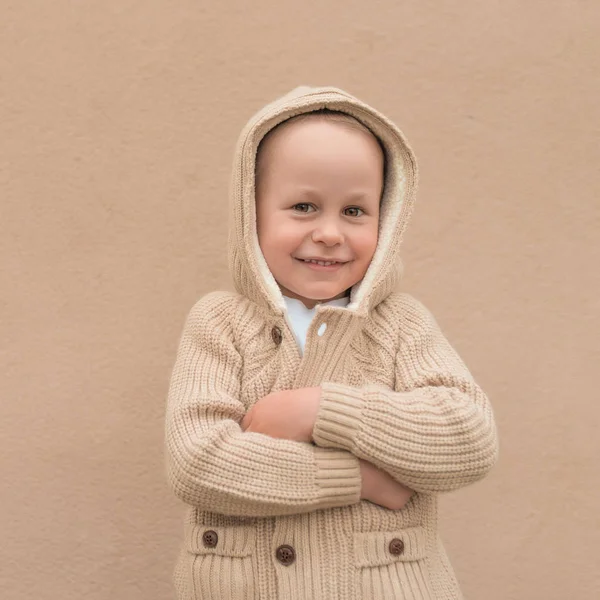 Malý chlapec 3-5 let, v béžovém svetru s kapucí, šťastně se usmívající, veselý a veselý. Pozadí zdi, podzim na ulici. Emoce potěšení a zábavy. — Stock fotografie