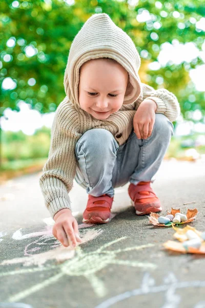 Маленький мальчик 3-5 лет, сидит на тротуаре и рисует желтое солнце, рисунки с красочными карандашами, творческие дети, воспитание и уход за детьми. В теплой одежде в осенний день на природе . — стоковое фото