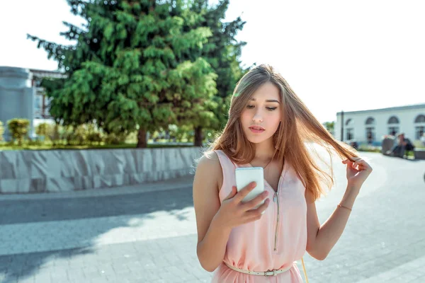 Seorang gadis bergaun merah muda, di musim panas di taman kota, membaca pesan di ponsel pintar, menonton video di ponsel, ruang kosong untuk menyalin teks . — Stok Foto