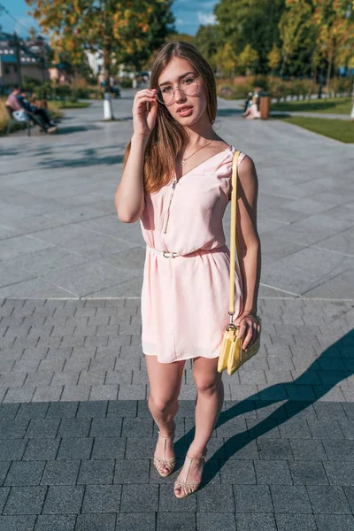 Chica en un vestido rosa, en el verano en un parque de la ciudad, posando con gafas, un bolso amarillo sobre su hombro. Hermosa mujer delgada y moderna . — Foto de Stock