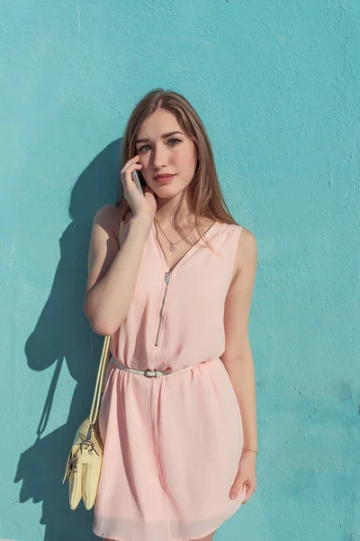 Chica en vestido rosa, en la pared de verano de fondo, hace llamadas telefónicas, escucha el mensaje de voz en la aplicación de Internet, un bolso amarillo. Hermosa mujer delgada y moderna . — Foto de Stock