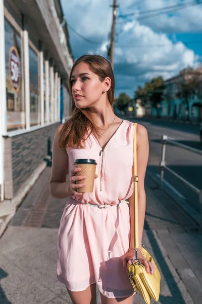 Gadis bergaun merah muda, latar belakang jalan musim panas, berdiri menunggu teman berpose, memegang cangkir dengan teh kopi, tas kuning. Cantik langsing dan wanita modern. Angka Tanned . — Stok Foto