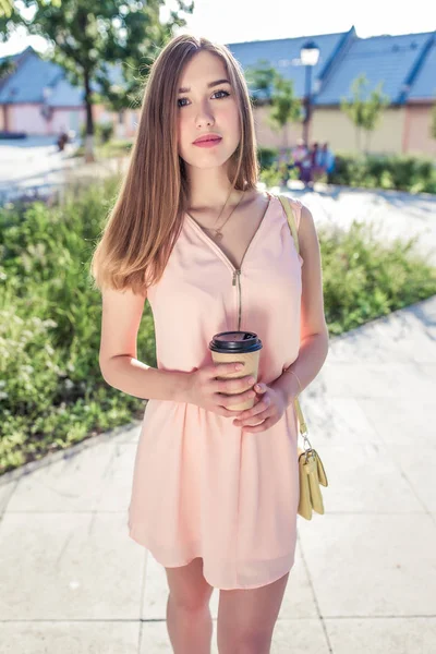 Dívka v růžových šatech, v létě v městském parku pózuje, v ruce drží sklenici čaje, žlutou kabelku. Moderní módní a krásná žena. Vyčiněná postava a dlouhé vlasy. — Stock fotografie