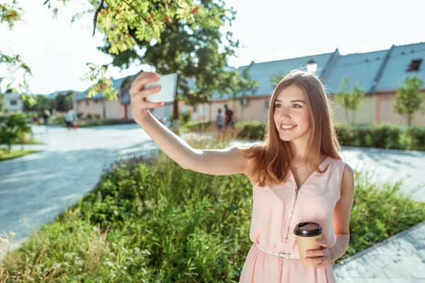 Dívka růžové šaty, letní město park obrázky na smartphone, selfie, drží šálek kávy čaj, usměje se šťastný, video hovor, on-line aplikace na internetu. Moderní módní krásná žena. Vyčiněné dlouhé vlasy. — Stock fotografie