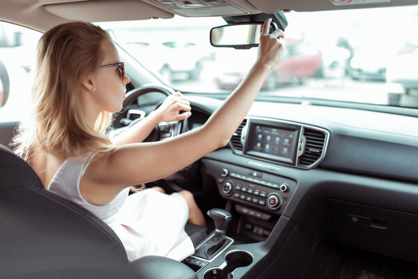 woman in car, adjusts and rearview mirror, driving safety in car, parking and rear traffic. Reverse parking in car. In summer in city, a pink dress. Automatic transmission.