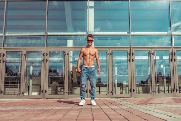 Young guy, athlete dancer, stands background of glass windows, trained happy smiling posing in summer in the city. Hip hop dancing. Jeans sunglasses. — Stok fotoğraf