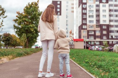 Mom walks with her son, autumn in the city, warm casual clothes, view from the back, background road grass and lawns.