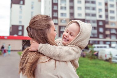 Woman mom holds a child in her arms, little boy son, autumn in the city, warm casual clothes, building background grass and lawns. Happy smiling have a rest at the weekend.