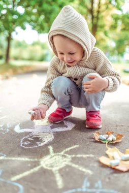 Little boy 3-5 years old, draws with crayons on asphalt drawing, creative idea art. Resting weekend, walk in autumn in park. Happy laughs and smiles. Warm casual wear, beige sweater with a hood.