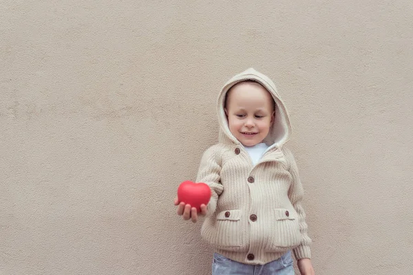 Menino de 3-5 anos, segura em sua mão brinquedo vermelho coração, regozija-se, fundo bege parede. Camisola quente com conceito de capuz de doação, presente, mães e dia dos namorados. Espaço livre para texto de cópia . — Fotografia de Stock