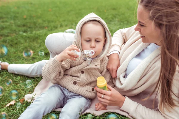 Máma žena si hraje s dítětem, chlapeček syn, podzimní ulice, foukání mýdlové bubliny, balónky, teplé ležérní oblečení, trávník pozadí. Emoce lásky starostlivý a relaxační víkend. — Stock fotografie