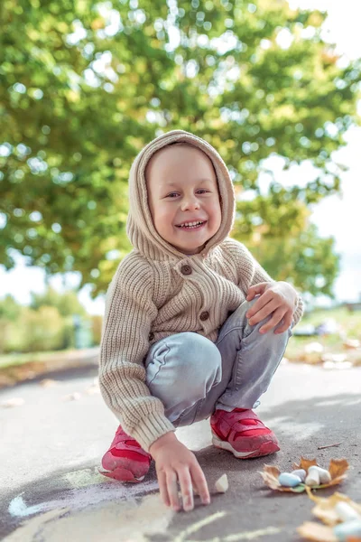 Маленький мальчик 3-5 лет, приседает, рисует цветными карандашами асфальт. Отдых в выходные, прогулка осенью в парке. Счастливый смех и улыбки. Теплая повседневная одежда, бежевый свитер с капюшоном . — стоковое фото