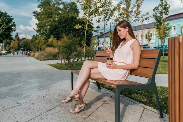 Chica en vestido rosa, sentado en el banco, en verano en el parque de la ciudad, sosteniendo el teléfono, taza de té de café. Lee mensajes SMS en las redes sociales, chat en línea en messenger. Espacio libre para copiar texto . —  Fotos de Stock