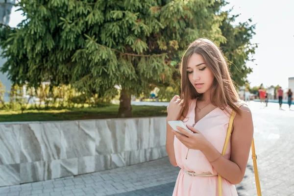Gadis bergaun pink, berdiri di musim panas di kota, tangan smartphone. Membaca pesan di aplikasi online, sms jaringan sosial. Ruang kosong untuk menyalin teks. Warna hijau latar belakang cemara . — Stok Foto