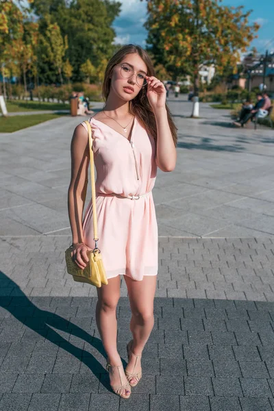 Hermosa chica en vestido rosa, se para en verano en la ciudad, ajusta las gafas, bolso amarillo, posando. Árboles de carretera de fondo pavimentando piedras. Emociones de relajación comodidad en un día soleado brillante . — Foto de Stock