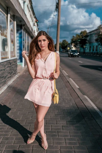 Mooi meisje roze jurk, staat zomerweg, poseren in volle groei, gele handtas. Wachtende vriendinnen. Emoties ontspanning comfort op heldere zonnige dag. Achtergrond straat winkels machine. — Stockfoto