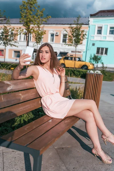 Chica en vestido rosa, sentado en el banco, verano en la ciudad, manos de teléfono inteligente. Fotografía en el teléfono, videollamada, aplicación en línea en Internet, grabación de mensajes de vídeo, autorretrato . — Foto de Stock