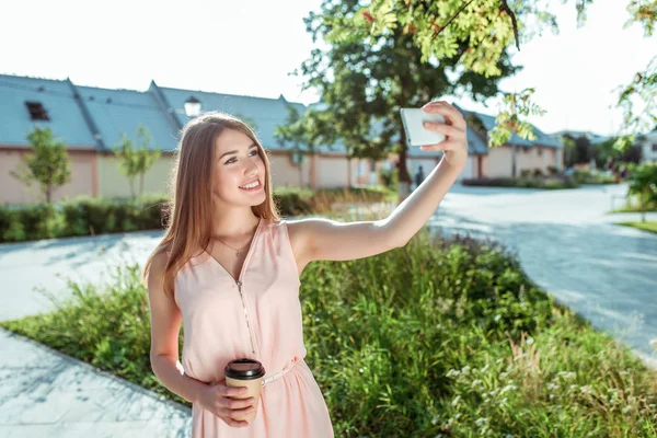 Šťastná dívka fotí sebe sama telefon, selfie na smartphonu, šálek kávy čaj v ruce, růžové šaty, v létě v městském parku, trávník stromy pozadí trávníku. Emoce štěstí, raduje se z relaxace. — Stock fotografie