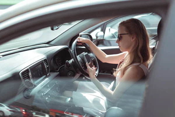 Donna in auto, seduta al volante di una macchina, legge e scrive messaggi nel telefono cellulare, nel parcheggio, nel traffico, in estate in auto. Ragazza in abito rosa, cambio automatico . — Foto Stock