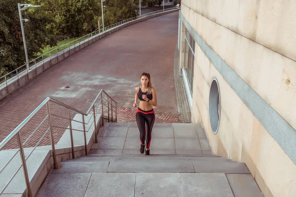 Красива спортивна дівчина біжить вранці, на літньому міському фоні сходи, мотивація способу життя. Спортивний одяг легінси зверху, таніфікована фігура. Вільне місце для тексту копіювання. Жінка в русі в стрибку . — стокове фото